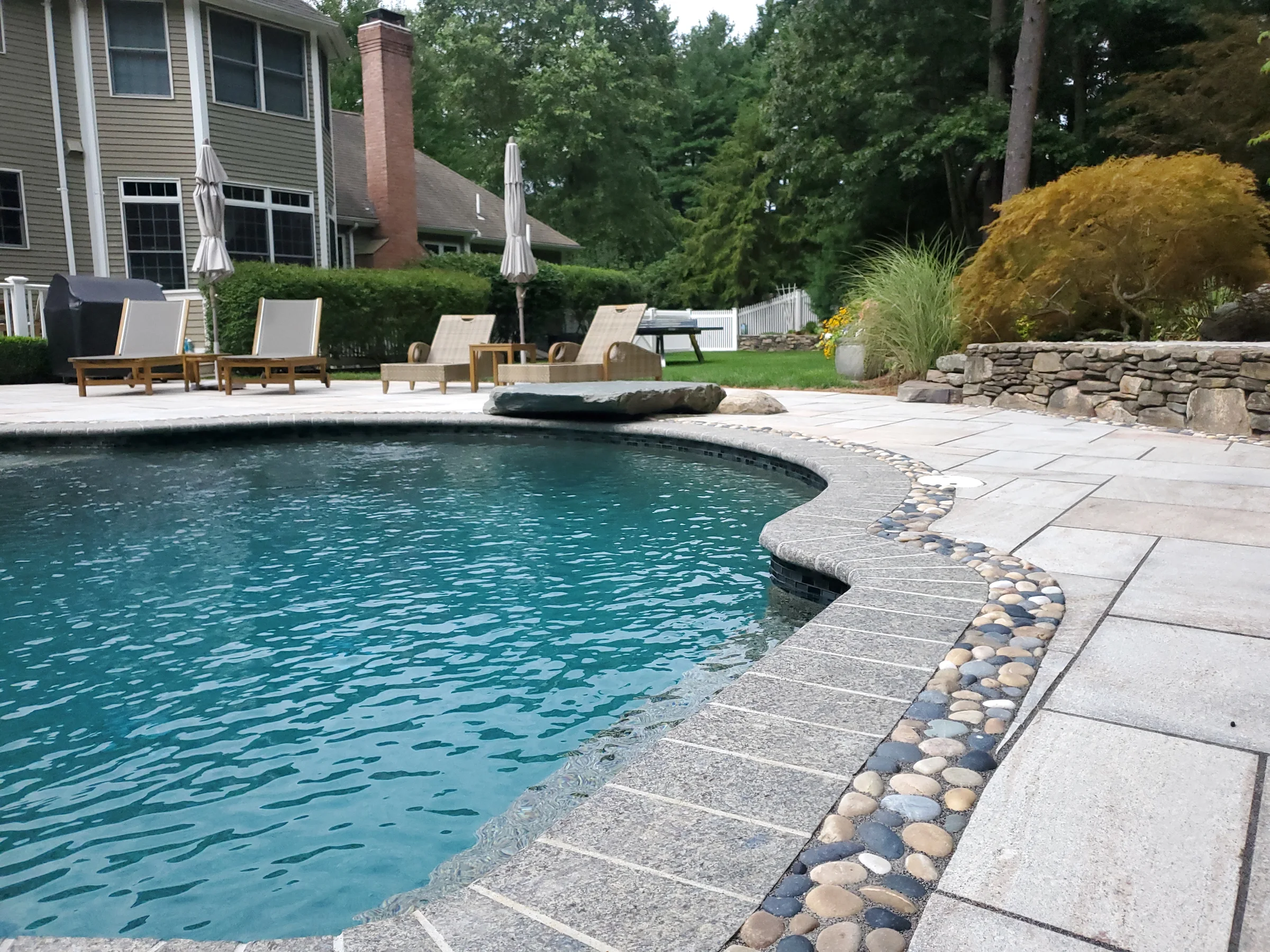 Curved swimming pool with pebble inlay, pale paving and a planted stone wall
